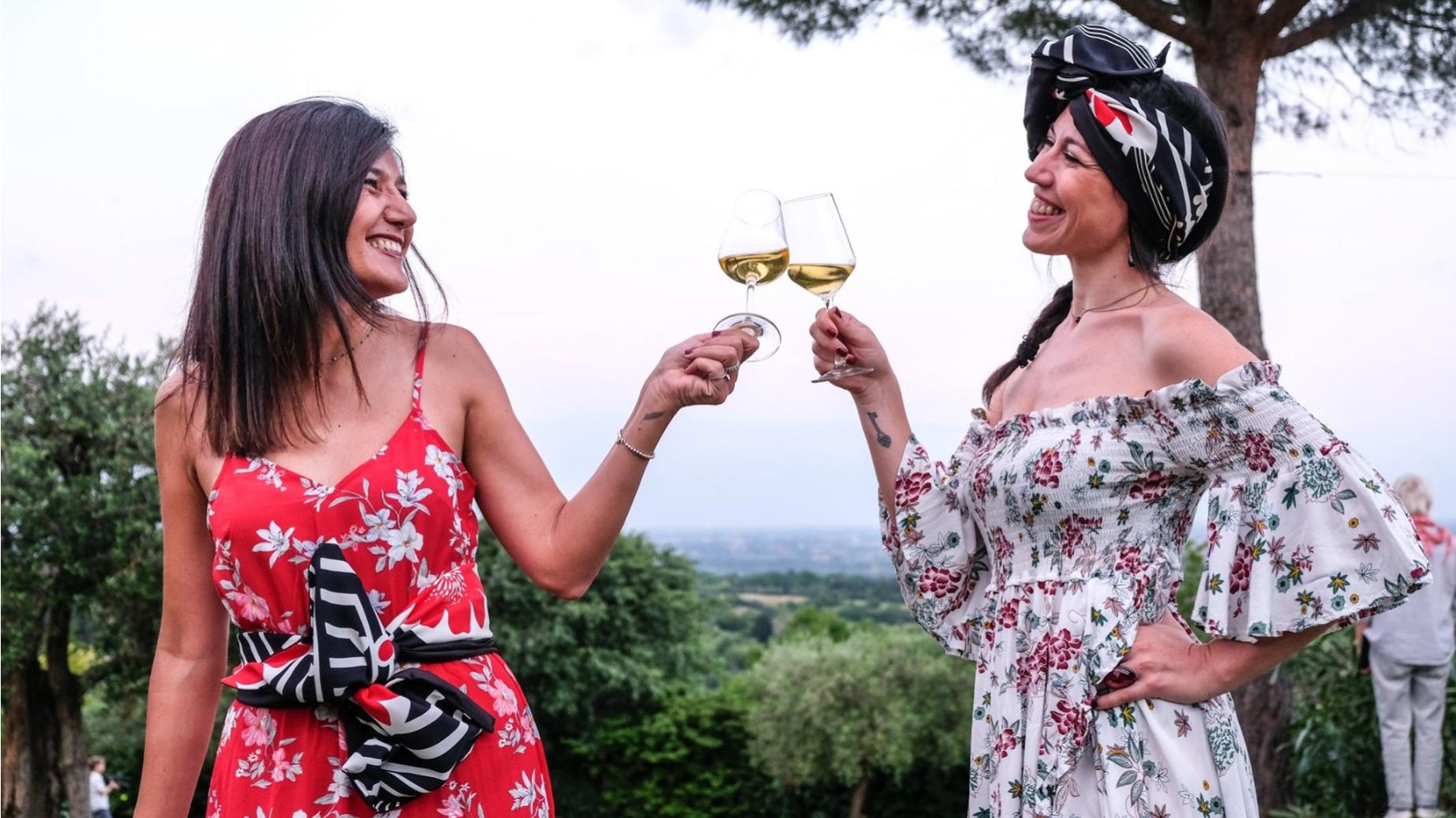 Due donne brindano con bicchieri di vino bianco in un ambiente all'aperto.