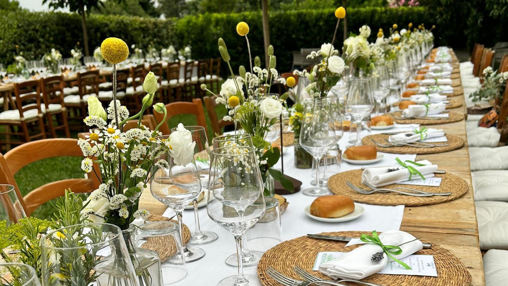 Tavola all'aperto elegantemente apparecchiata con fiori, bicchieri e tovaglioli legati con nastro verde.