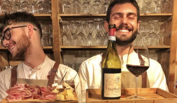 Due ragazzi sorridenti con vino e tagliere di salumi e formaggi in una cantina.