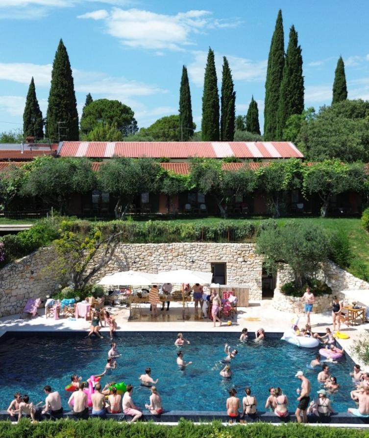 Piscina affollata in un giardino con cipressi e un edificio rustico sullo sfondo.